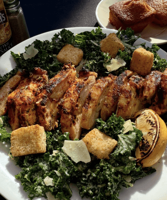 A plate of sliced grilled chicken breast served over a bed of kale Caesar salad with croutons, shaved parmesan, and a charred lemon wedge from La Bola. Sunlight highlights the fresh ingredients.